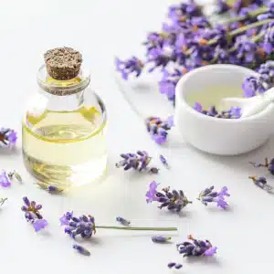 Une bouteille d'huile en verre avec un bouchon en liège, entourée de brins de lavande. Un bol blanc contenant de l'huile et une cuillère se trouvent à l'arrière-plan.